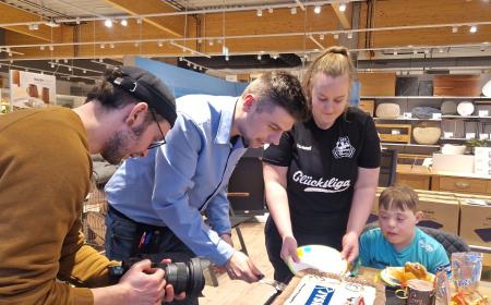 Ein Mann in JYSK-Hemd schneidet eine Torte an. Eine Frau mit Pferdeschwanz hält einen Teller. Ein Mann mit Mütze und Brille filmt die Szene mit einer Kamera.